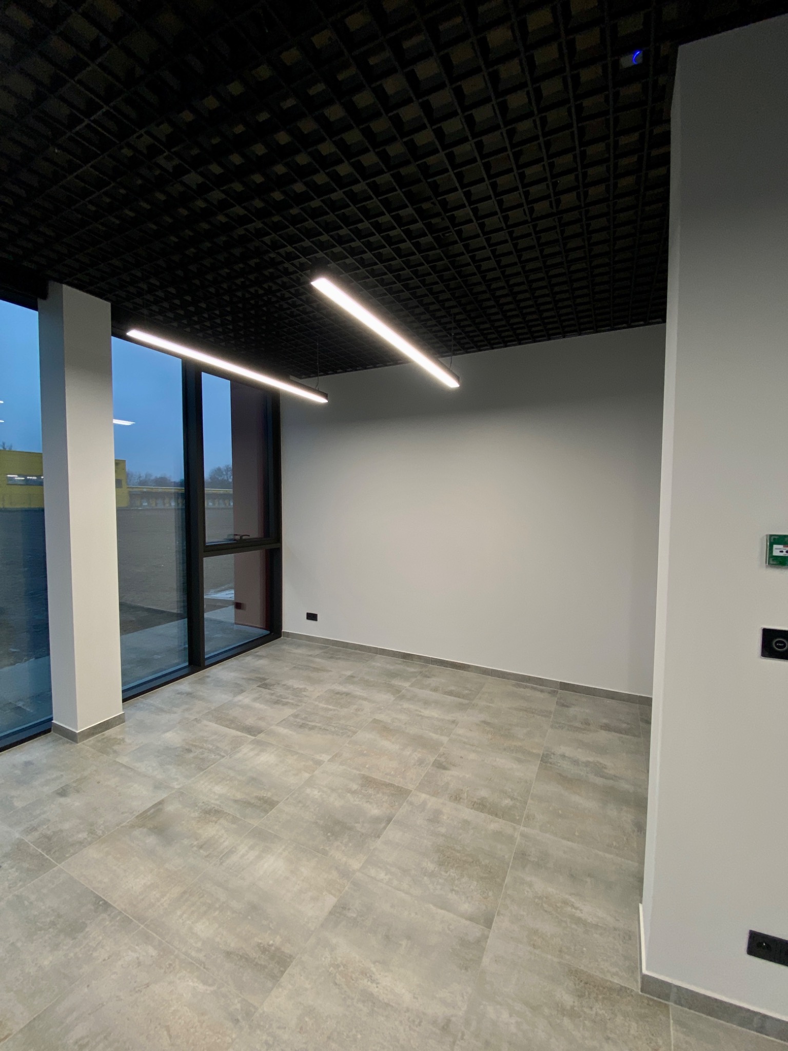 Nowoczesne wnętrze biurowe z szarą podłogą, białymi ścianami i czarnym sufitem rastrowym. Długie lampy LED oświetlają przestrzeń. Widoczne duże okna z widokiem na zewnątrz.