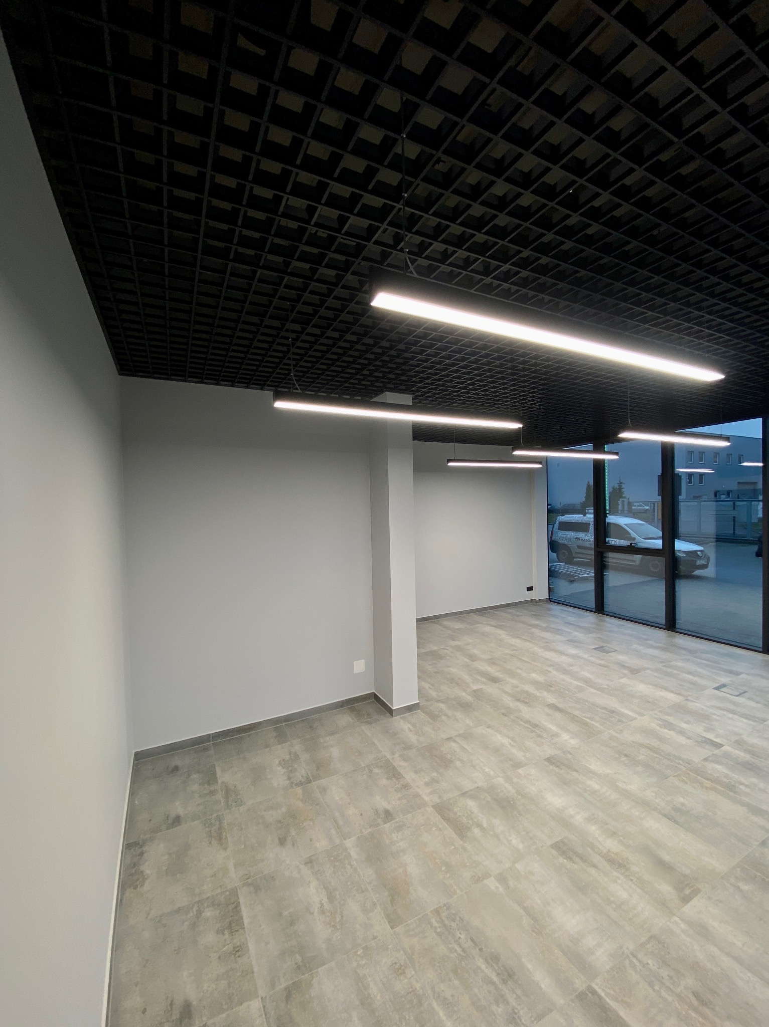 Wnętrze biurowe po remoncie: szare ściany, podłoga z płytek imitujących drewno, czarny sufit rastrowy z podłużnymi lampami LED. Widok na zewnątrz przez szklane drzwi.