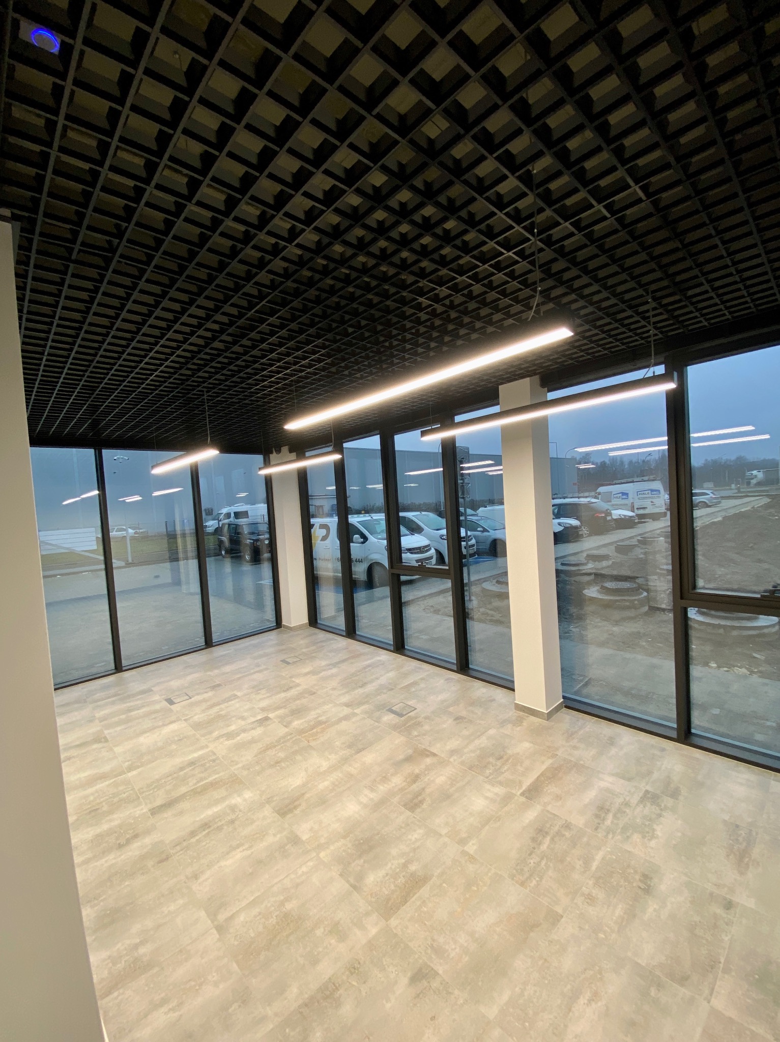 Nowoczesne wnętrze biurowe z wykończoną podłogą z płytek, przeszklonymi ścianami i podwieszanym sufitem w kratkę z oświetleniem liniowym.