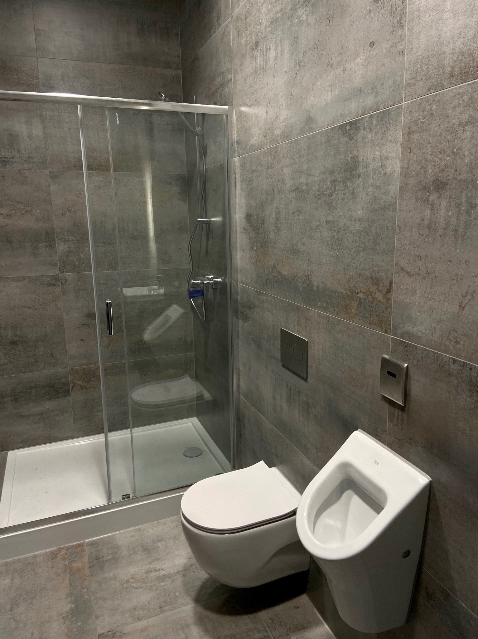 Nowoczesna łazienka z kabiną prysznicową, toaletą i pisuarzem. Szare płytki imitujące beton na ścianach i podłodze. Minimalistyczne akcesoria łazienkowe.