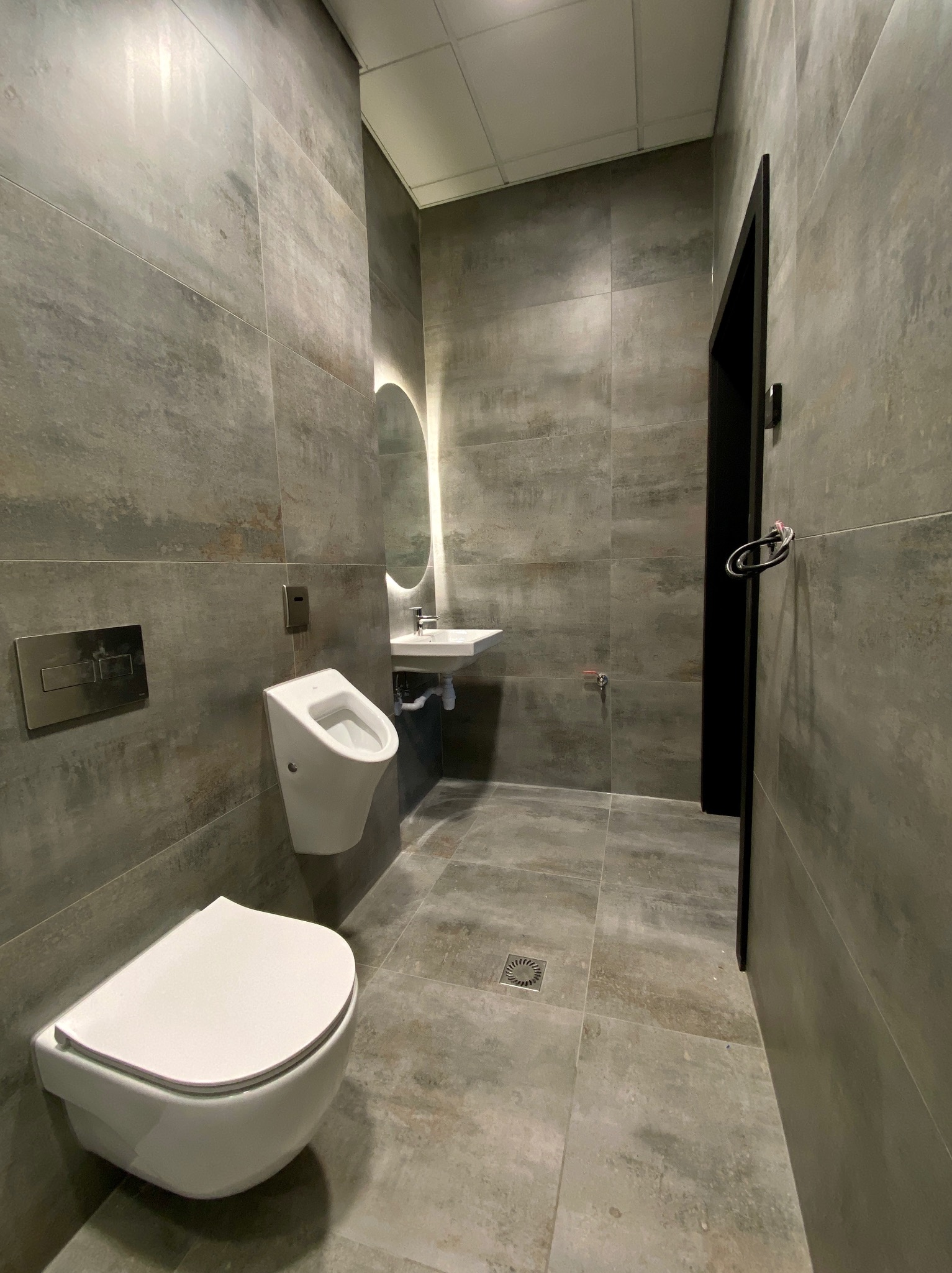 Wykończona łazienka w Bochni z szarymi płytkami, wiszącą toaletą i pisuar, okrągłym lustrem z podświetleniem i czarną futryną drzwi. Nowoczesny design.