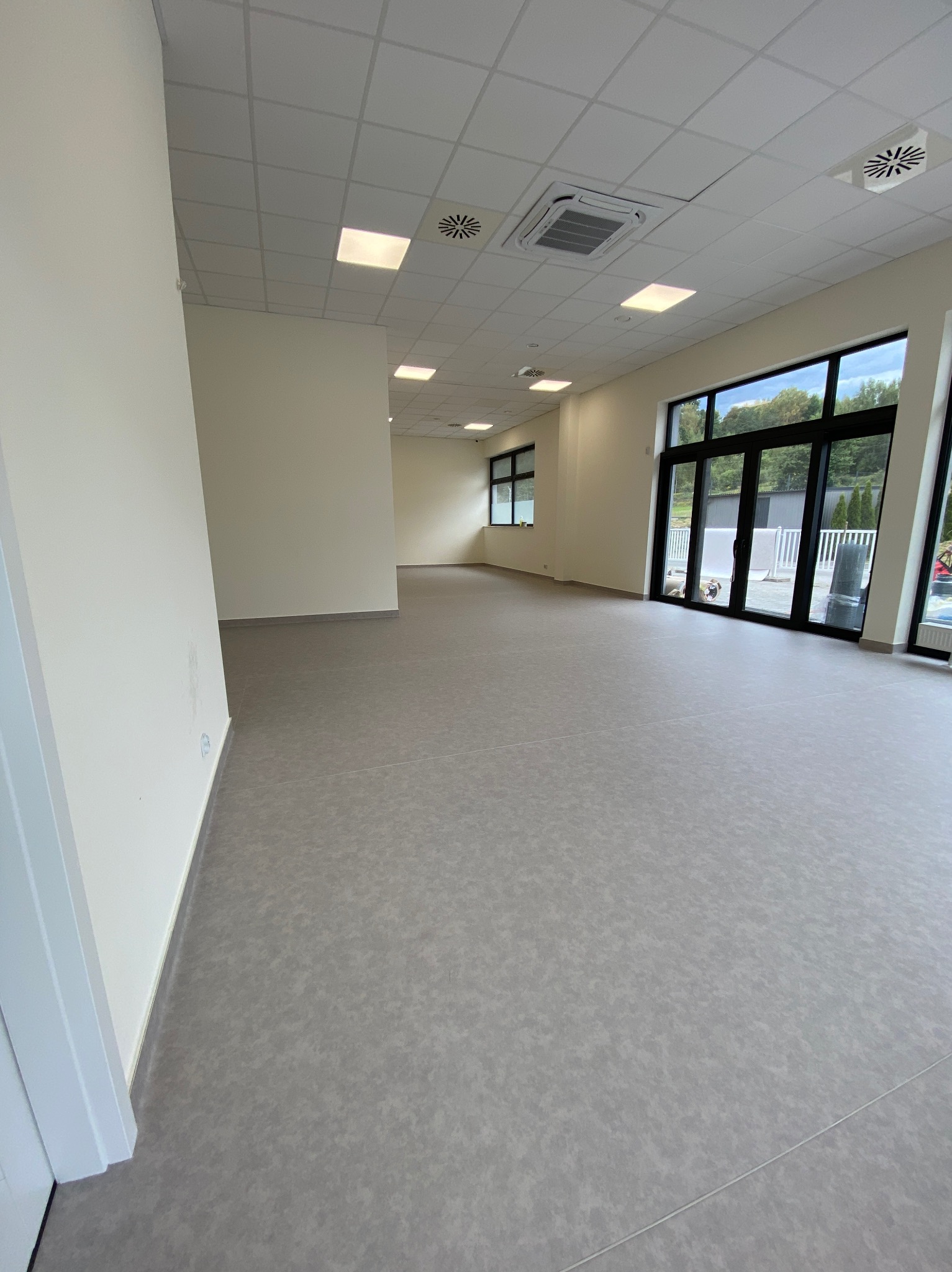 Jasne, przestronne wnętrze po remoncie z szarą podłogą, białymi ścianami i dużymi oknami. Widoczne elementy wentylacji na suficie. Minimalistyczny design.