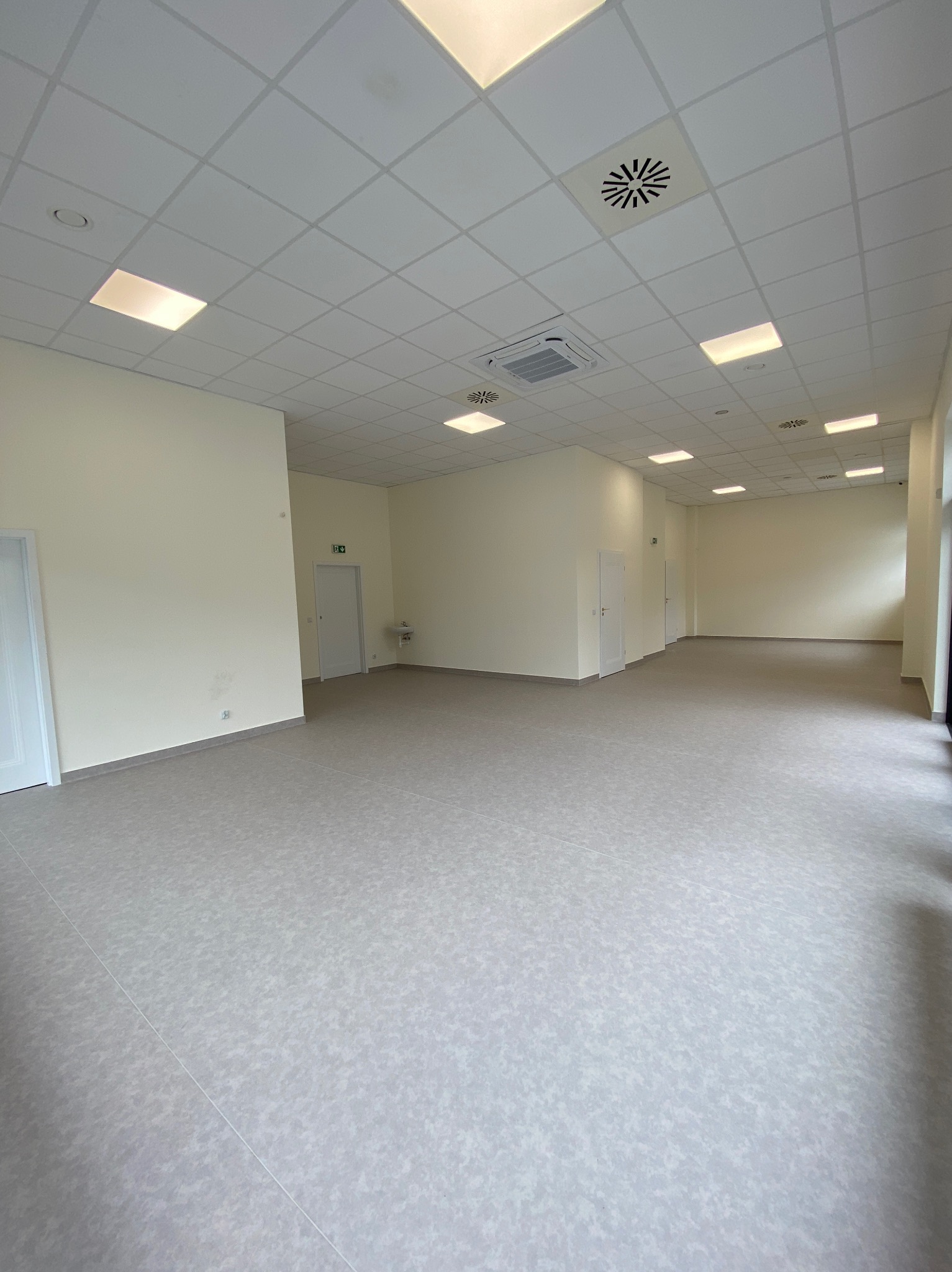 Jasne, przestronne wnętrze z podwieszanym sufitem kasetonowym, oświetleniem LED i szarą podłogą. Widoczne drzwi i elementy wentylacji.