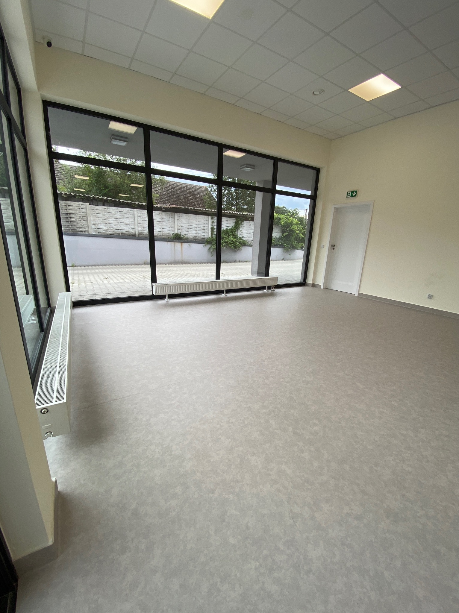 Jasne, przestronne wnętrze z dużymi oknami i szarą podłogą. Widoczne grzejniki pod oknami i białe ściany. Minimalistyczny design, sufit kasetonowy z oświetleniem.