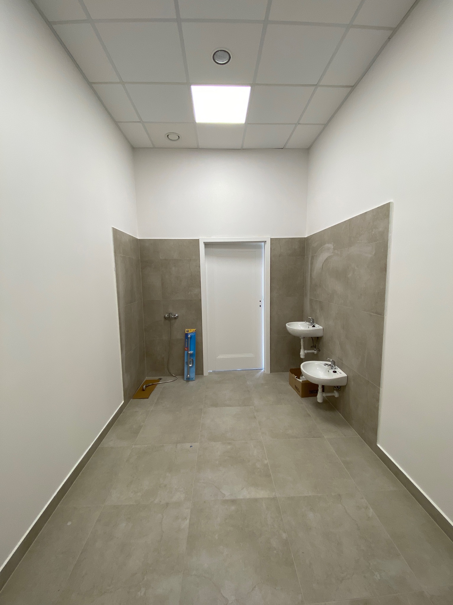 Wykończona łazienka w odcieniach szarości i bieli. Dwie umywalki, płytki na ścianach do połowy wysokości, białe ściany powyżej, sufit kasetonowy z oświetleniem.