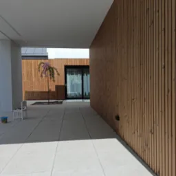 Nowoczesny taras z jasną, betonową posadzką i ścianą wykończoną pionowymi panelami z jasnego drewna, małe drzewko w donicy, cień na posadzce.