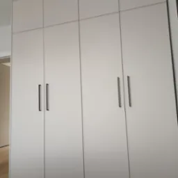 Zamknięta, czterodrzwiowa szafa wnękowa z matowymi, jasnymi frontami i pionowymi, czarnymi uchwytami, umieszczona we wnętrzu mieszkania.