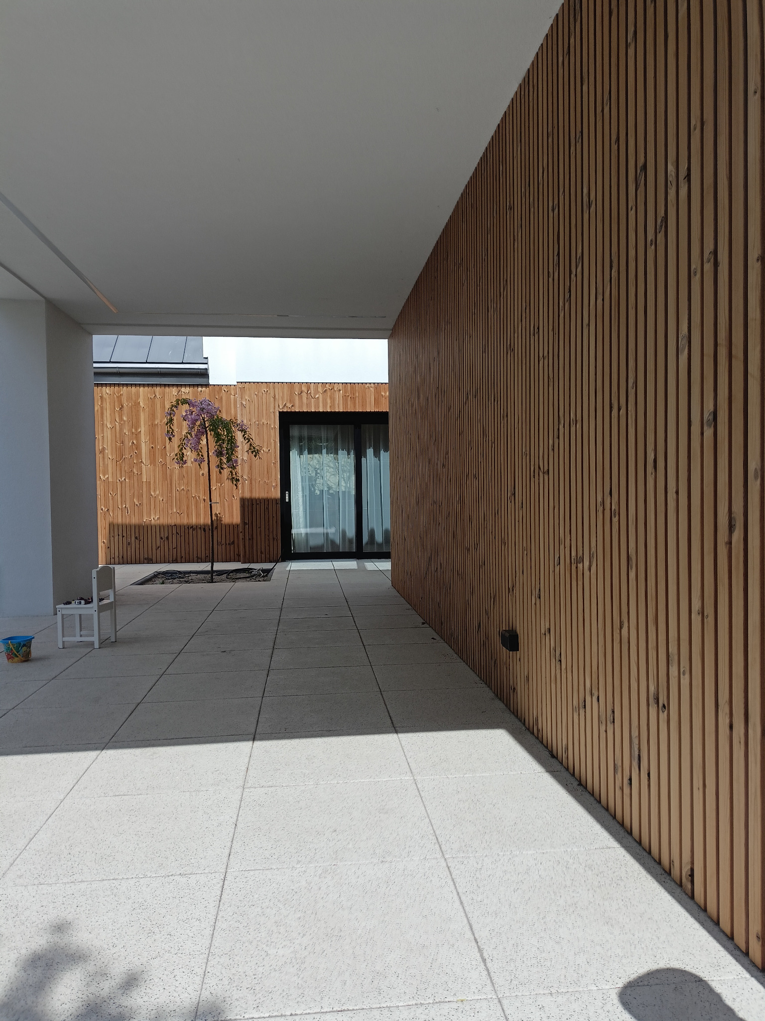 Nowoczesny taras z jasną, betonową posadzką i ścianą wykończoną pionowymi panelami z jasnego drewna, małe drzewko w donicy, cień na posadzce.