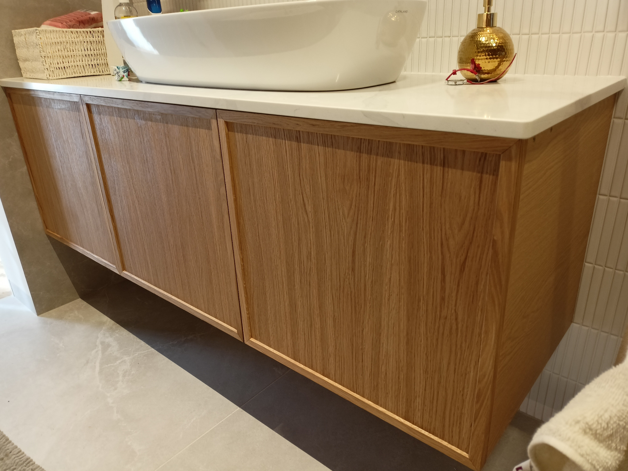 Nowoczesna szafka łazienkowa z jasnego drewna z blatem z białego konglomeratu, na którym stoi owalna umywalka i złota butelka z dozownikiem, widok z dołu.