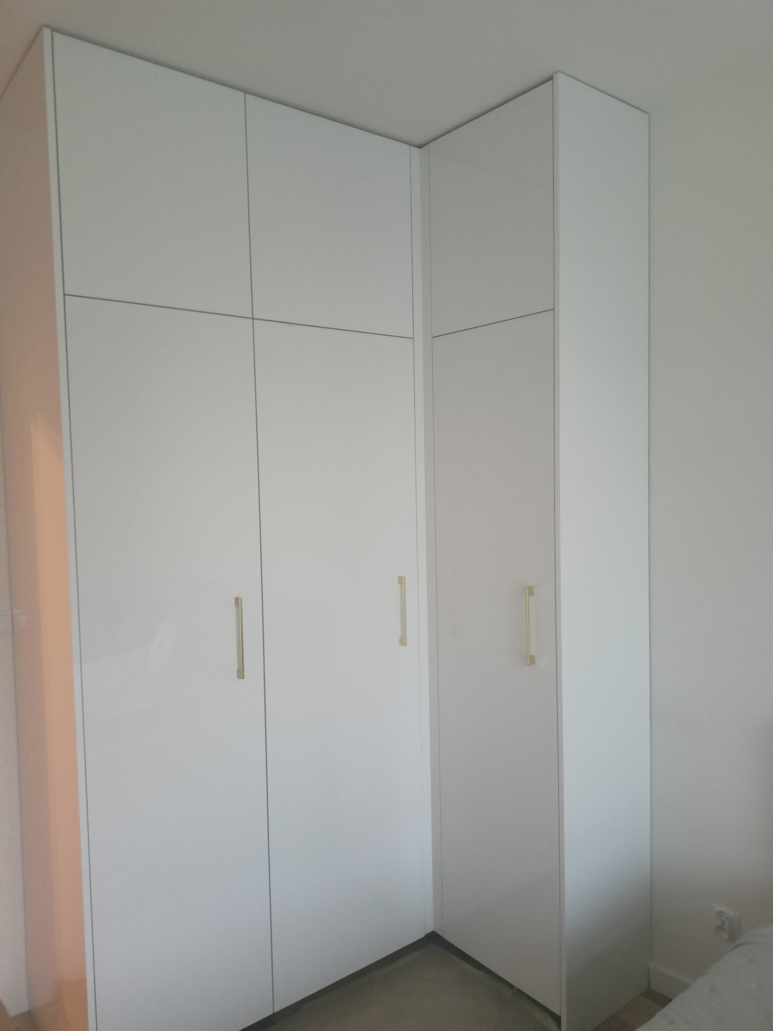 Biała, narożna szafa z połyskiem i złotymi uchwytami, dopasowana do kąta pokoju, widoczna z bliska.