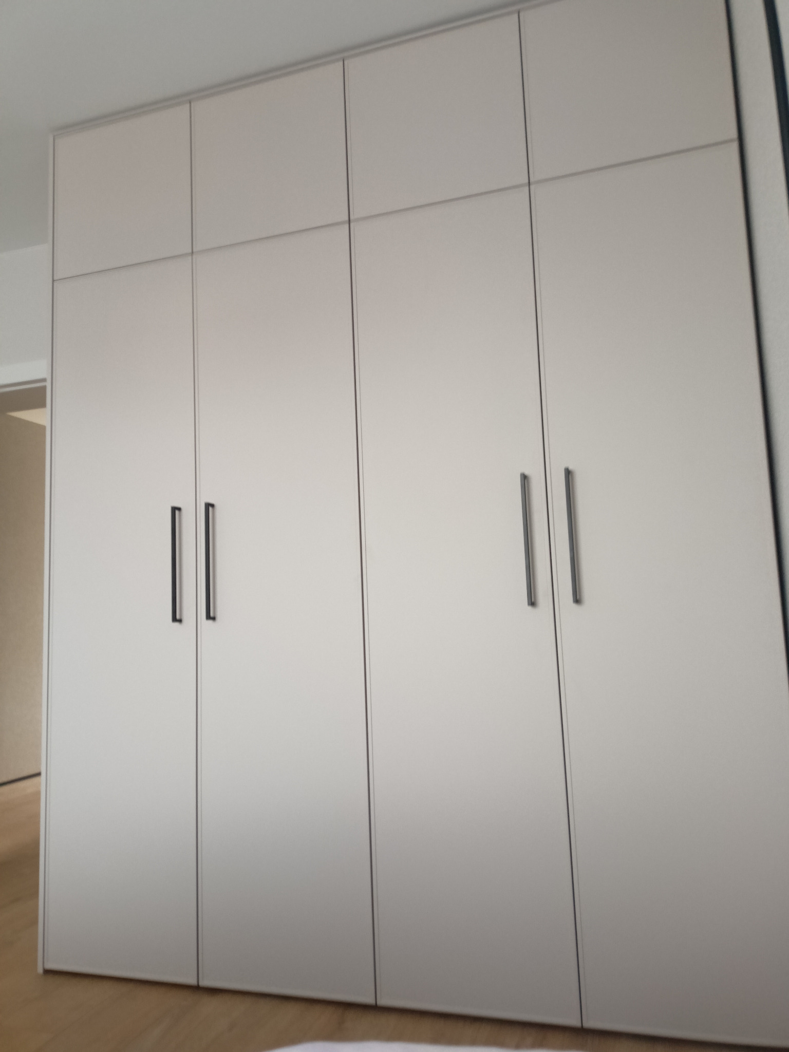 Zamknięta, czterodrzwiowa szafa wnękowa z matowymi, jasnymi frontami i pionowymi, czarnymi uchwytami, umieszczona we wnętrzu mieszkania.