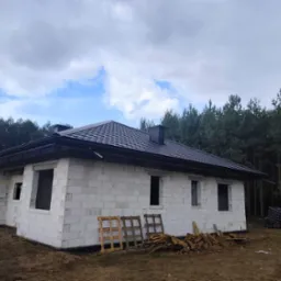 Dom w stanie surowym zamkniętym, z czarnym dachem, wykonany z białych bloczków. Widoczne palety i drewno. Tło stanowią drzewa i pochmurne niebo.