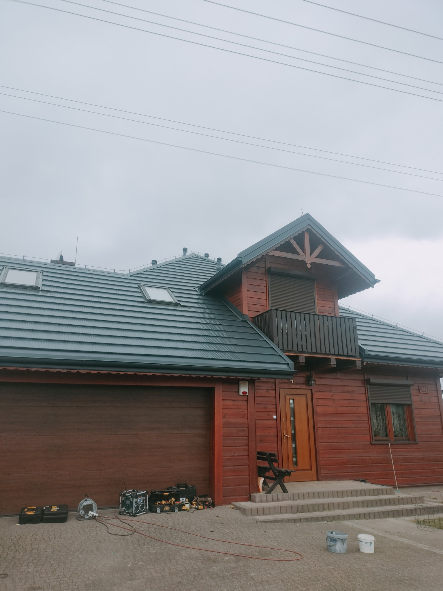 Dom z nowym, grafitowym dachem z dachówki metalowej, widoczne okna dachowe, drewniana elewacja, narzędzia i puszki z farbą przed garażem.