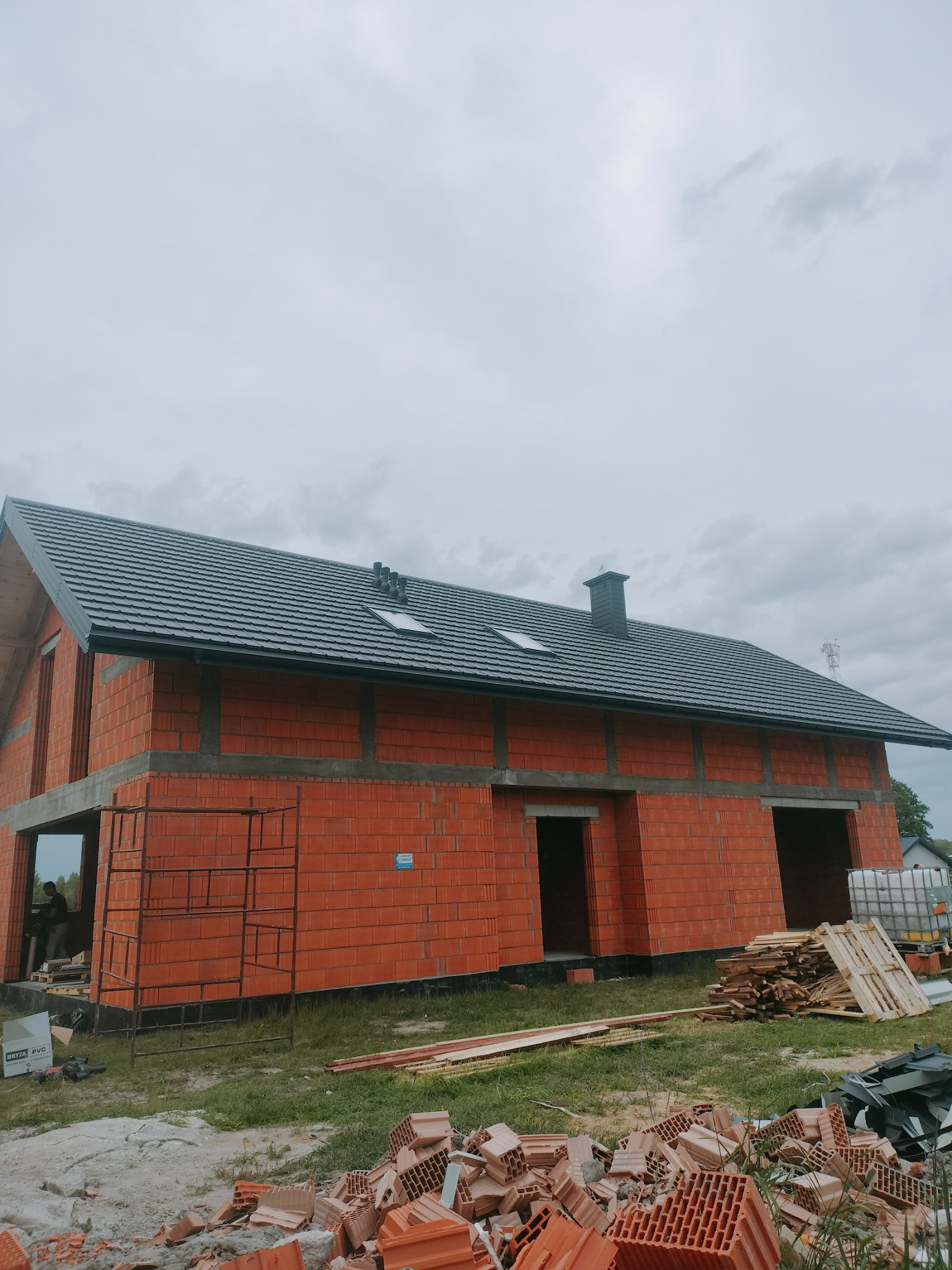 Ceglany dom w trakcie budowy z ciemnym dachem i kominem. Widoczne rusztowania, palety z materiałami budowlanymi i stos cegieł. Pochmurne niebo w tle.