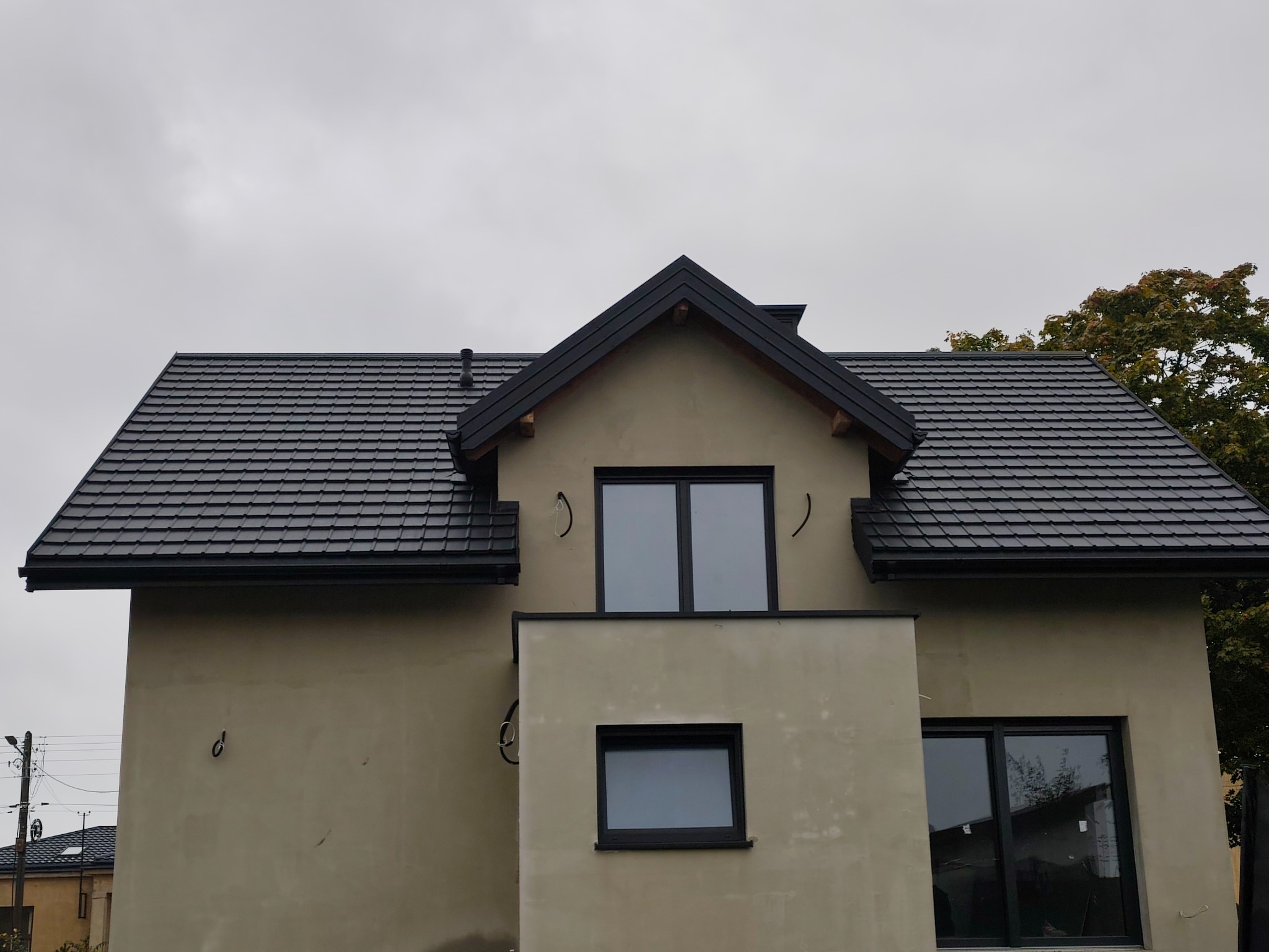 Dom z nowym, czarnym dachem z dachówki ceramicznej, widoczny z dołu, z elementami elewacji w surowym stanie. Na ścianie zamocowane metalowe uchwyty.