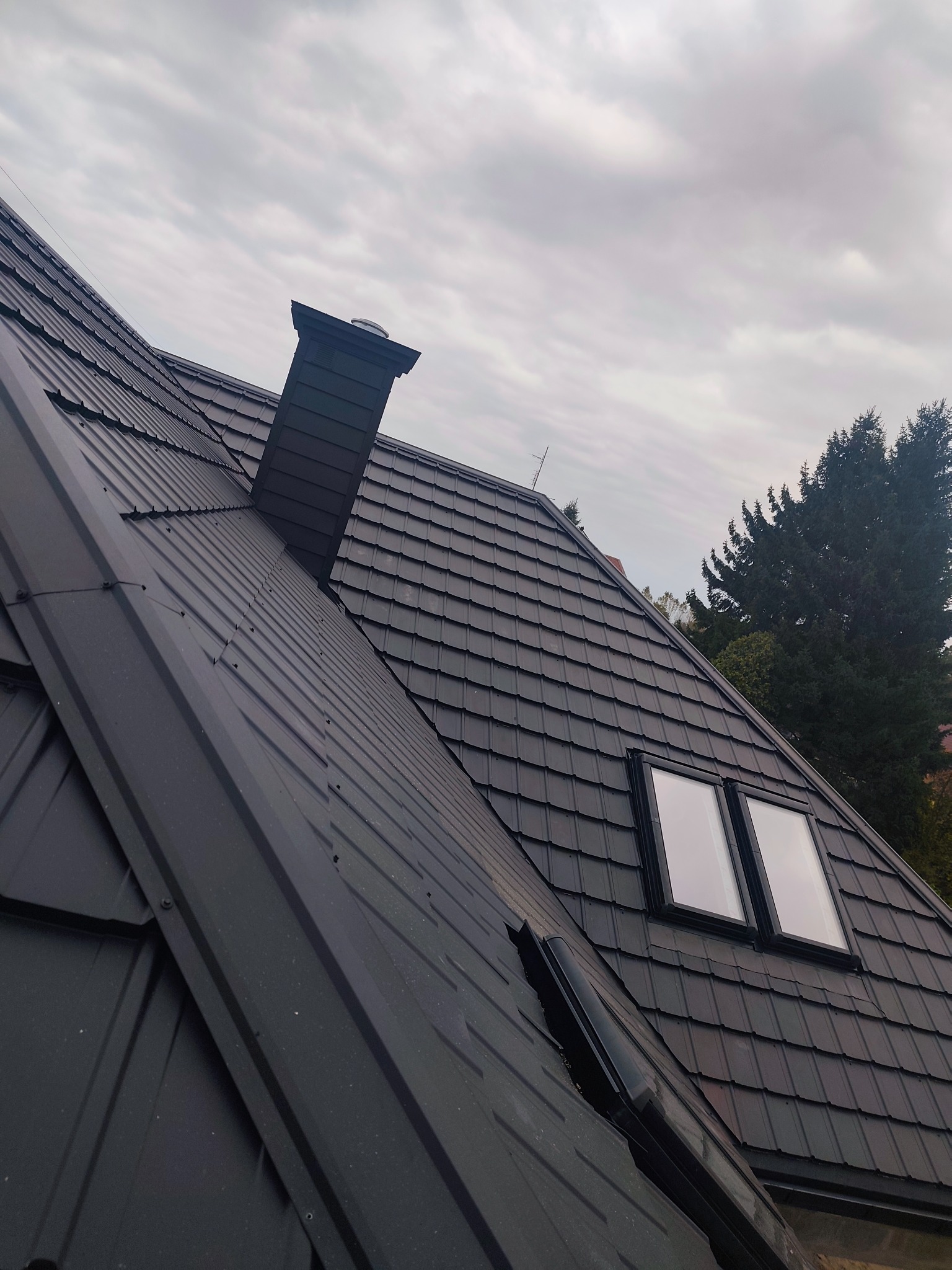 Czarny dach z blachy trapezowej z kominem i dwoma oknami dachowymi, widok z dołu na tle pochmurnego nieba i zieleni drzew.