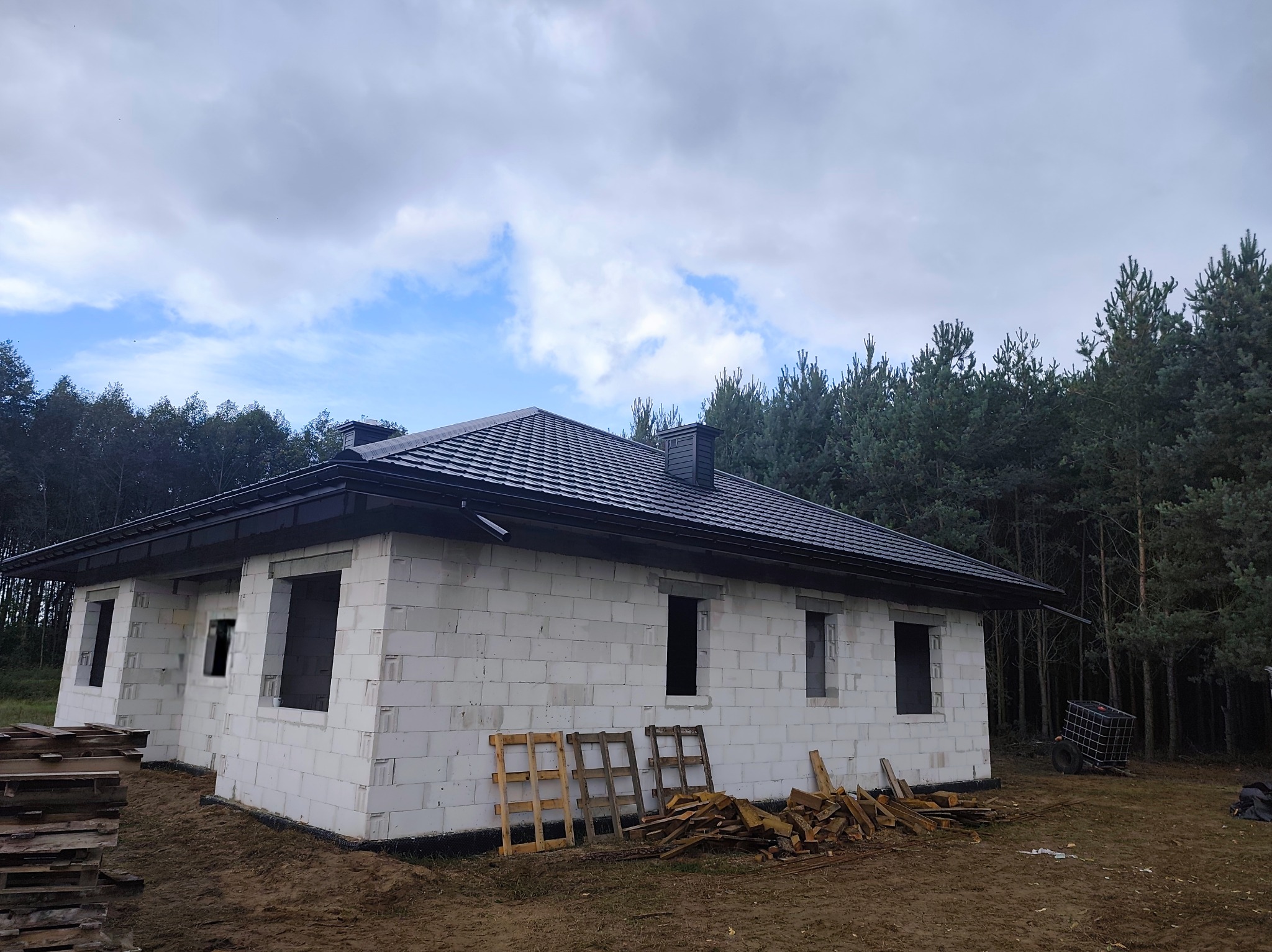 Dom w stanie surowym zamkniętym, z czarnym dachem, wykonany z białych bloczków. Widoczne palety i drewno. Tło stanowią drzewa i pochmurne niebo.
