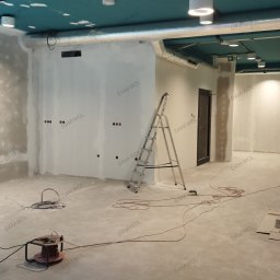DAM-WOL renovation - Wnętrze w trakcie prac wykończeniowych: ściany z widocznymi miejscami po szpachlowaniu, aluminiowa drabina, lampa budowlana na statywie, zwinięte kable, oraz fragment sufitu w kolorze turkusowym.
