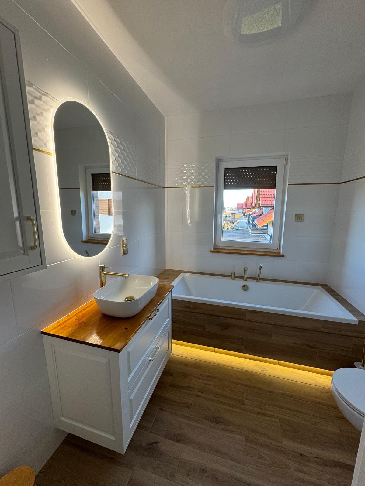 Nowoczesna łazienka na poddaszu z biało-drewnianymi akcentami: podświetlane lustro, umywalka na drewnianym blacie, wanna z drewnianą obudową i podświetleniem LED.