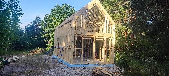 Surowa konstrukcja domu w trakcie budowy, widoczne drewniane ramy ścian i dachu na fundamencie, otoczone zielenią drzew i krzewów, z niebieskimi rurami widocznymi na zewnątrz.