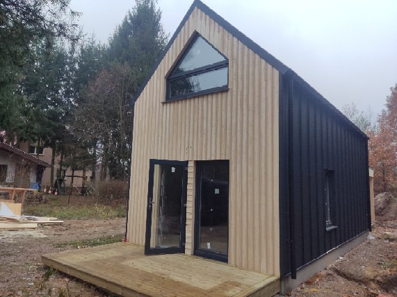 Nowoczesny dom o dwukolorowej elewacji z pionowym deskowaniem, z drewnianym tarasem i trójkątnym oknem na szczycie, w otoczeniu drzew i zabudowań.