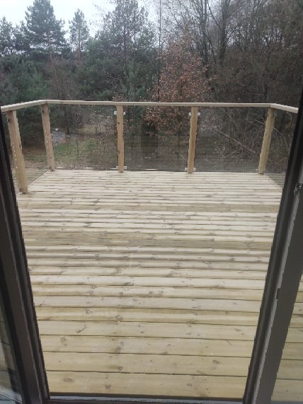 Drewniany taras z balustradą ze szklanym wypełnieniem, widok z wnętrza domu przez otwarte drzwi balkonowe, w tle drzewa.