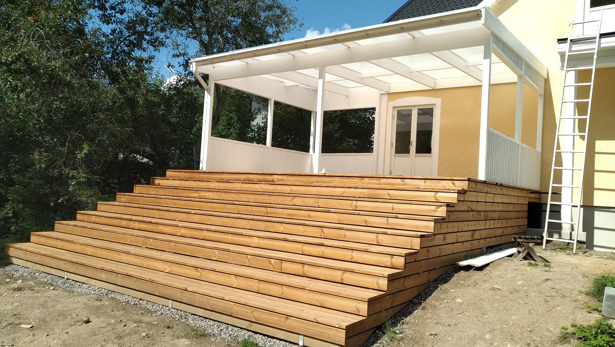 Drewniane schody prowadzące do zadaszonego tarasu przy domu z żółtą elewacją, drabina oparta o ścianę, widok z dołu.