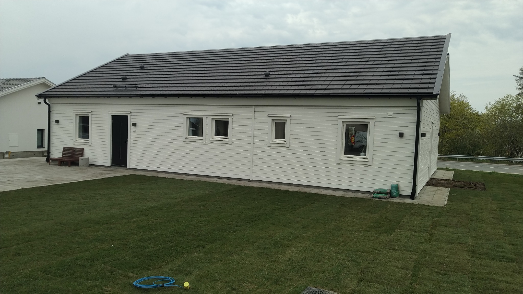 Nowoczesny, parterowy dom z białymi panelami elewacyjnymi i grafitowym dachem, otoczony świeżo założonym trawnikiem, widok z boku na tle pochmurnego nieba.