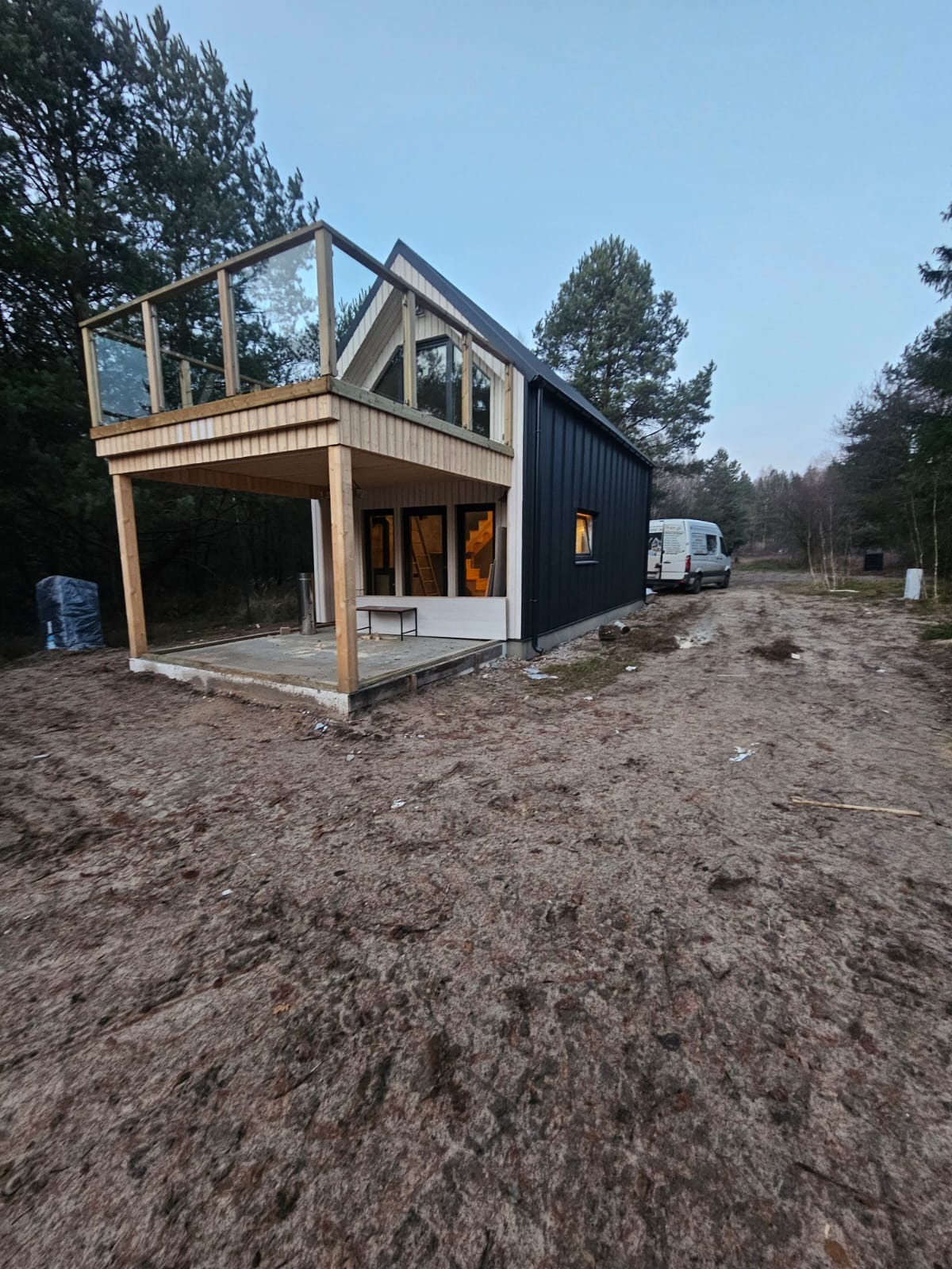 Nowoczesny dom z drewnianą elewacją i balkonem ze szklaną balustradą, częściowo wykończony, z widocznym samochodem dostawczym w tle.
