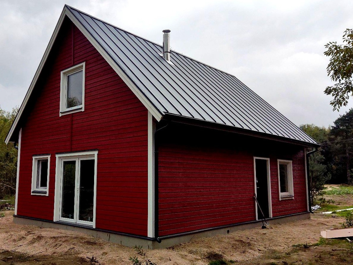 Czerwony dom z drewnianą elewacją i metalowym dachem na tle pochmurnego nieba, widok z boku.