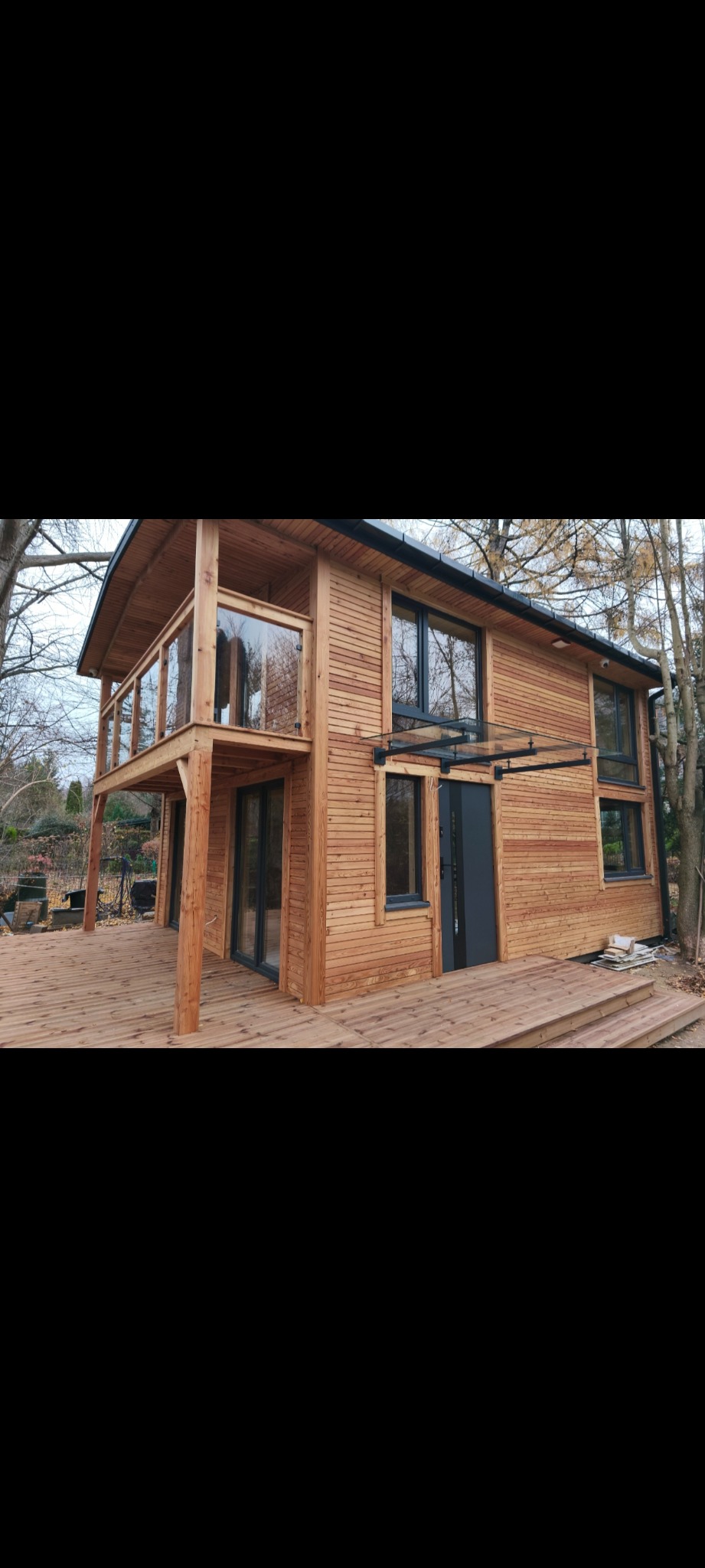 Dwukondygnacyjny dom szkieletowy z drewnianą elewacją, balkonem i szklanym zadaszeniem nad wejściem. Widok z zewnątrz, w tle drzewa.