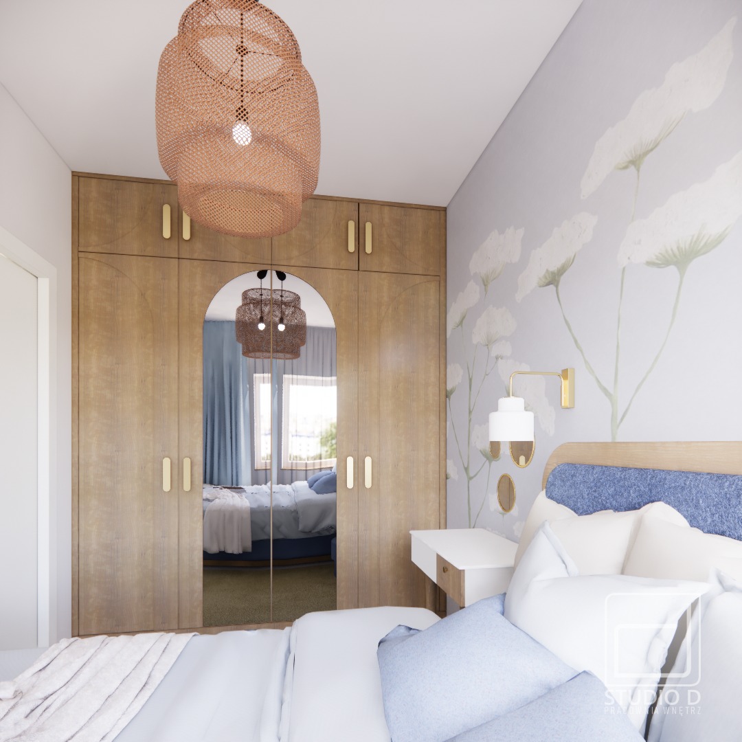 Jasna sypialnia z drewnianą szafą z lustrem, lampą z plecionki i tapetą w kwiaty. Widok na łóżko i okno w odbiciu lustra. Spokojny, przytulny wystrój.