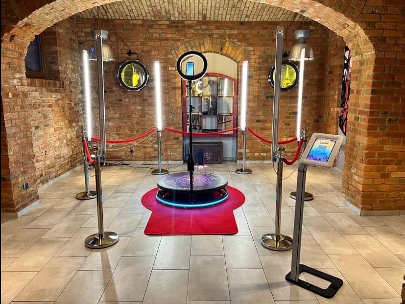 Wnętrze z ceglanymi ścianami, gdzie znajduje się okrągła platforma z podświetleniem LED, otoczona czerwonym dywanem i oświetlona przez cztery lampy LED na statywach. Na platformie umieszczony jest...