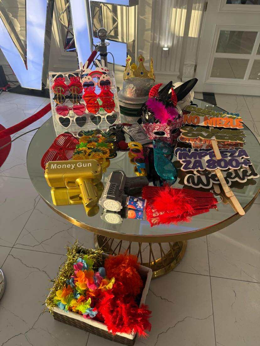 Różnorodne rekwizyty na szklanym stoliku: okulary, korony, maski, rekwizyty z napisami i pistolety na pieniądze. Elementy dekoracji imprezowych.