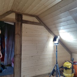 ANDRZEJ SŁABY MODRZEW - Poddasze wykończone jasną boazerią, z widoczną drewnianą konstrukcją nośną pomalowaną na ciemny brąz, oświetlone lampą budowlaną na statywie, z odkurzaczem przemysłowym w rogu i częściowo widoczną...