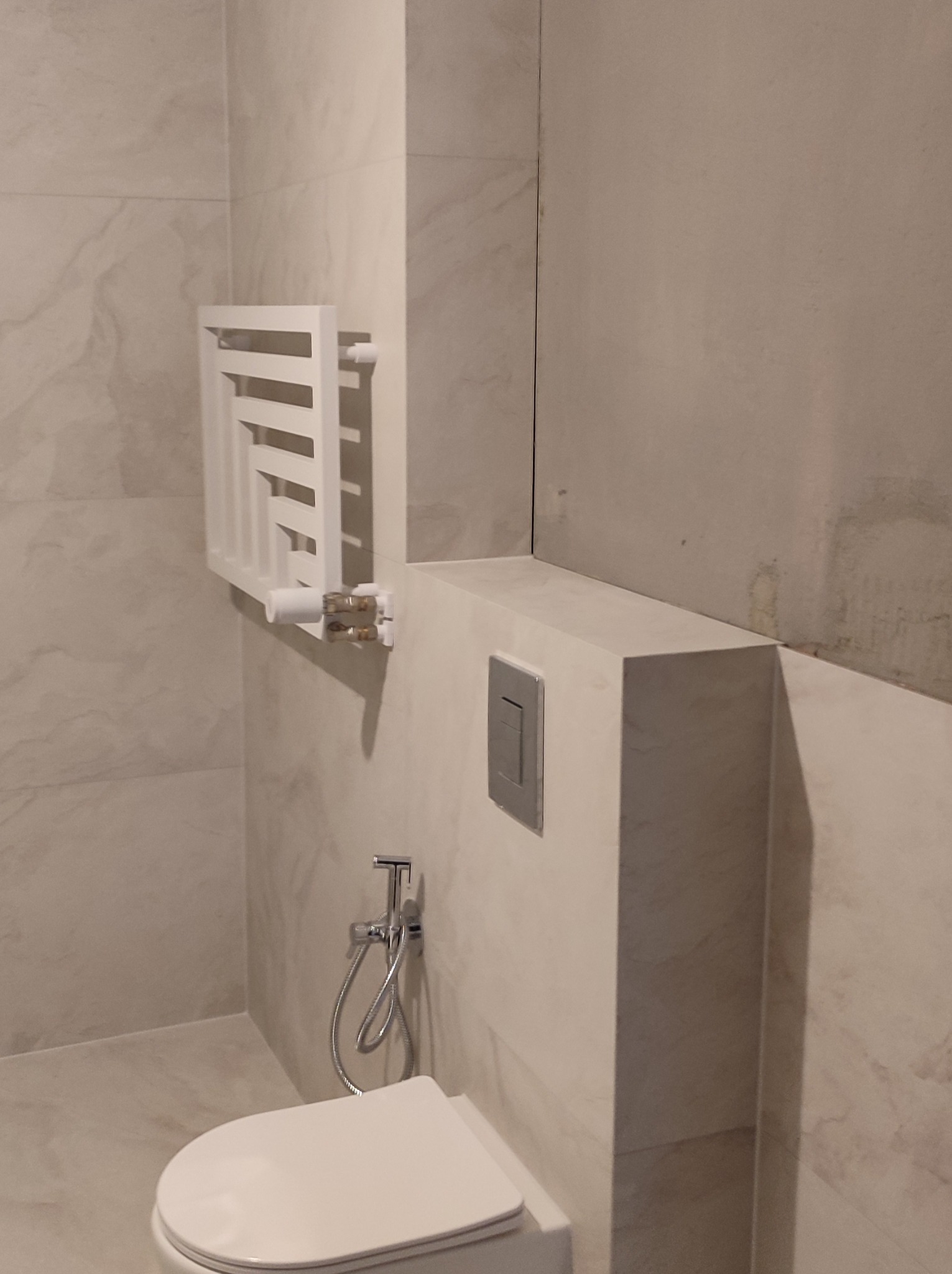 Bidetka obok toalety, biały grzejnik drabinkowy na ścianie z marmurowym wzorem, szary przycisk spłuczki na zabudowie.