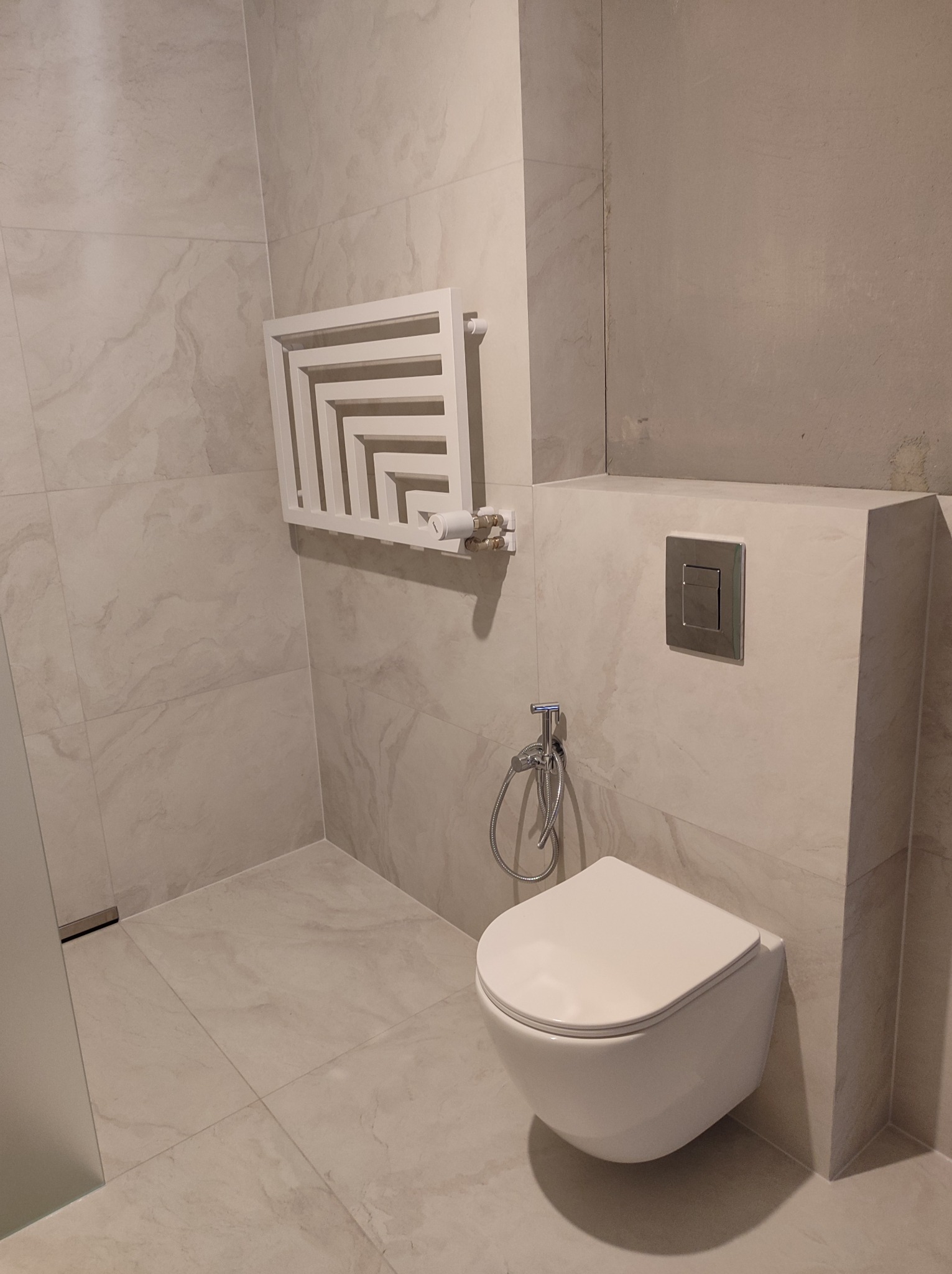 Nowoczesna toaleta podwieszana z bidetką i geometrycznym grzejnikiem drabinkowym na tle płytek imitujących marmur w odcieniach beżu i szarości.