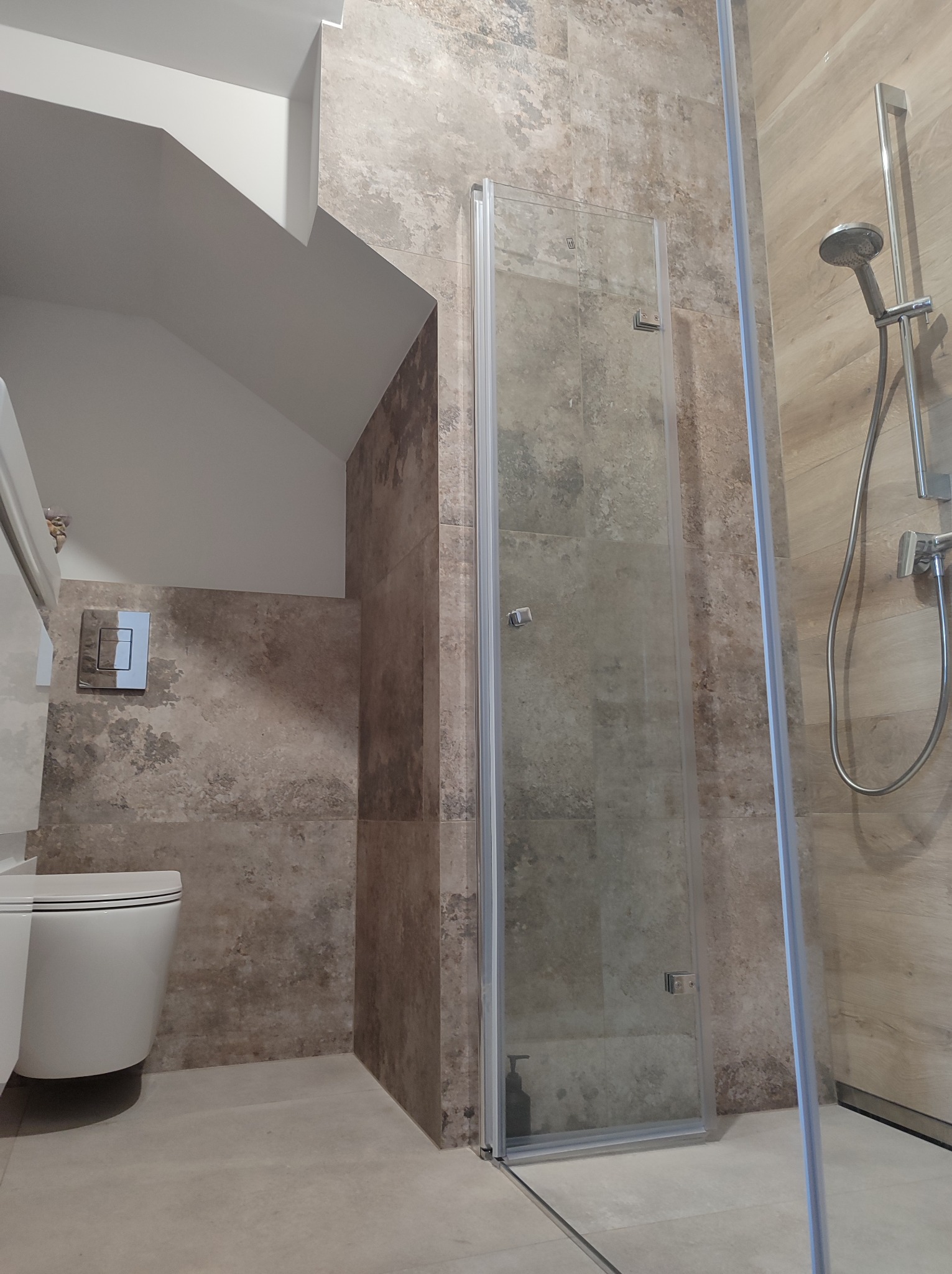 Nowoczesna łazienka z beżowymi płytkami imitującymi kamień, kabina prysznicowa z transparentnymi drzwiami, wisząca toaleta z chromowanym przyciskiem spłukującym i zestaw prysznicowy.