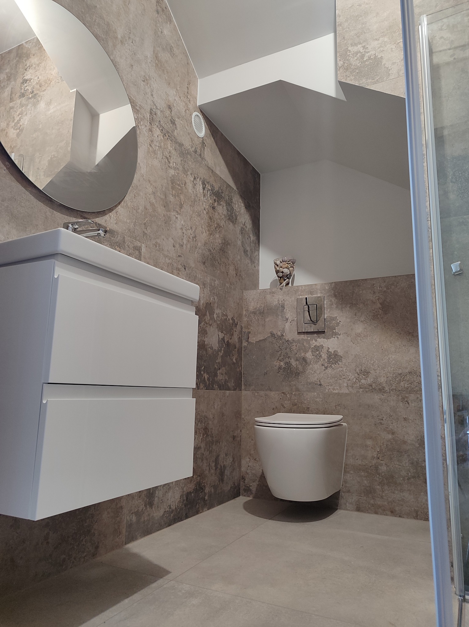 Nowoczesna łazienka z umywalką na szafce, wiszącą toaletą i dużym owalnym lustrem, wykończona płytkami imitującymi beton w odcieniach szarości i brązu, z dekoracyjnym wazonem z kamieniami na półce.