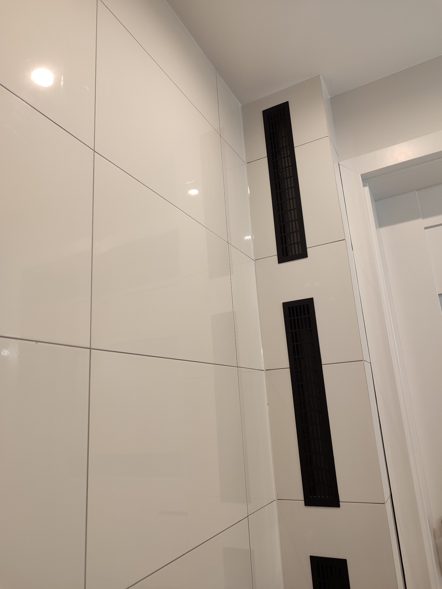 Białe, błyszczące płytki ceramiczne ułożone w łazience w Łodzi, z zamontowanymi czarnymi kratkami wentylacyjnymi, widoczne fragmenty białych drzwi.