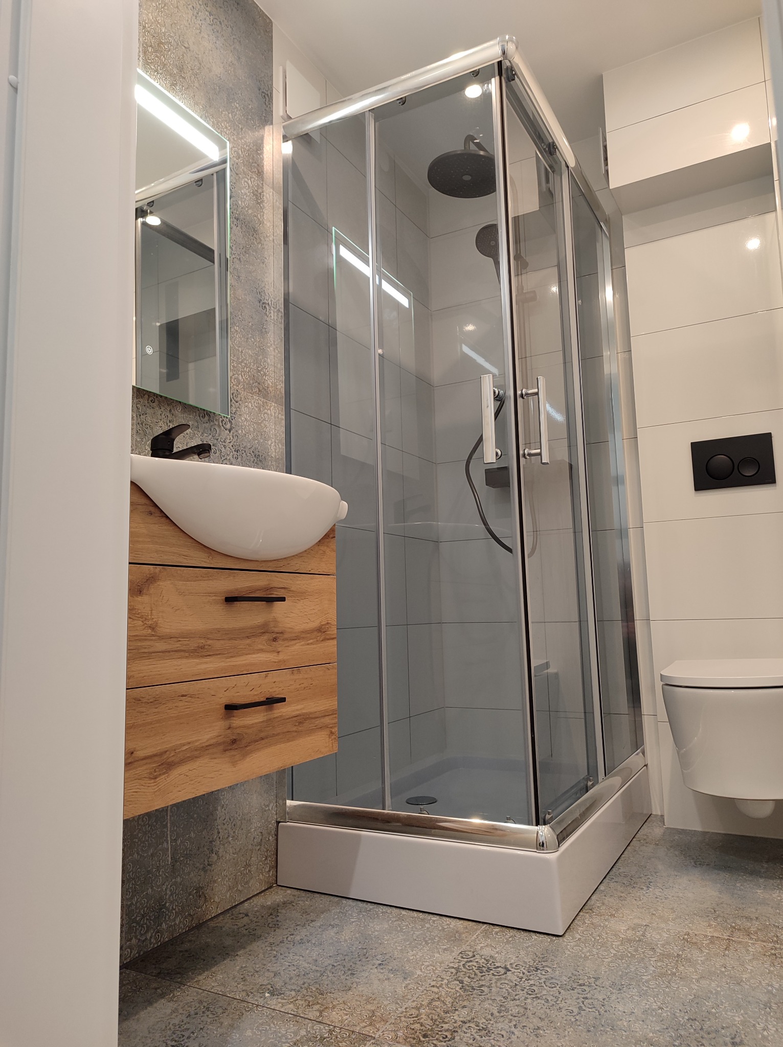 Nowoczesna łazienka z kabiną prysznicową, umywalką na drewnianej szafce i podwieszaną toaletą, wykończona w szaro-białej kolorystyce z dekoracyjnymi płytkami podłogowymi.