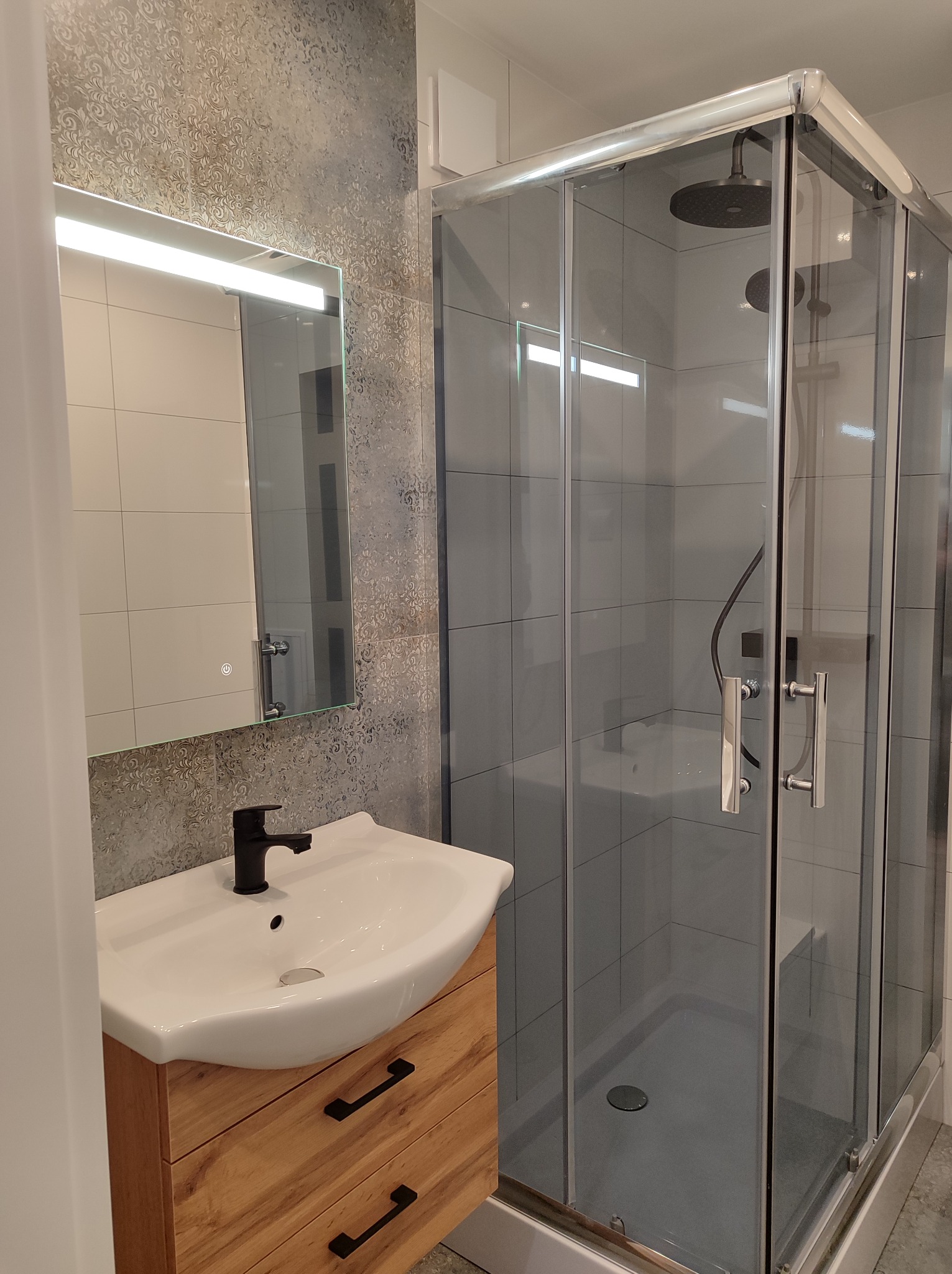 Nowoczesna łazienka z kabiną prysznicową, umywalką na drewnianej szafce i podświetlanym lustrem na tle dekoracyjnej ściany.