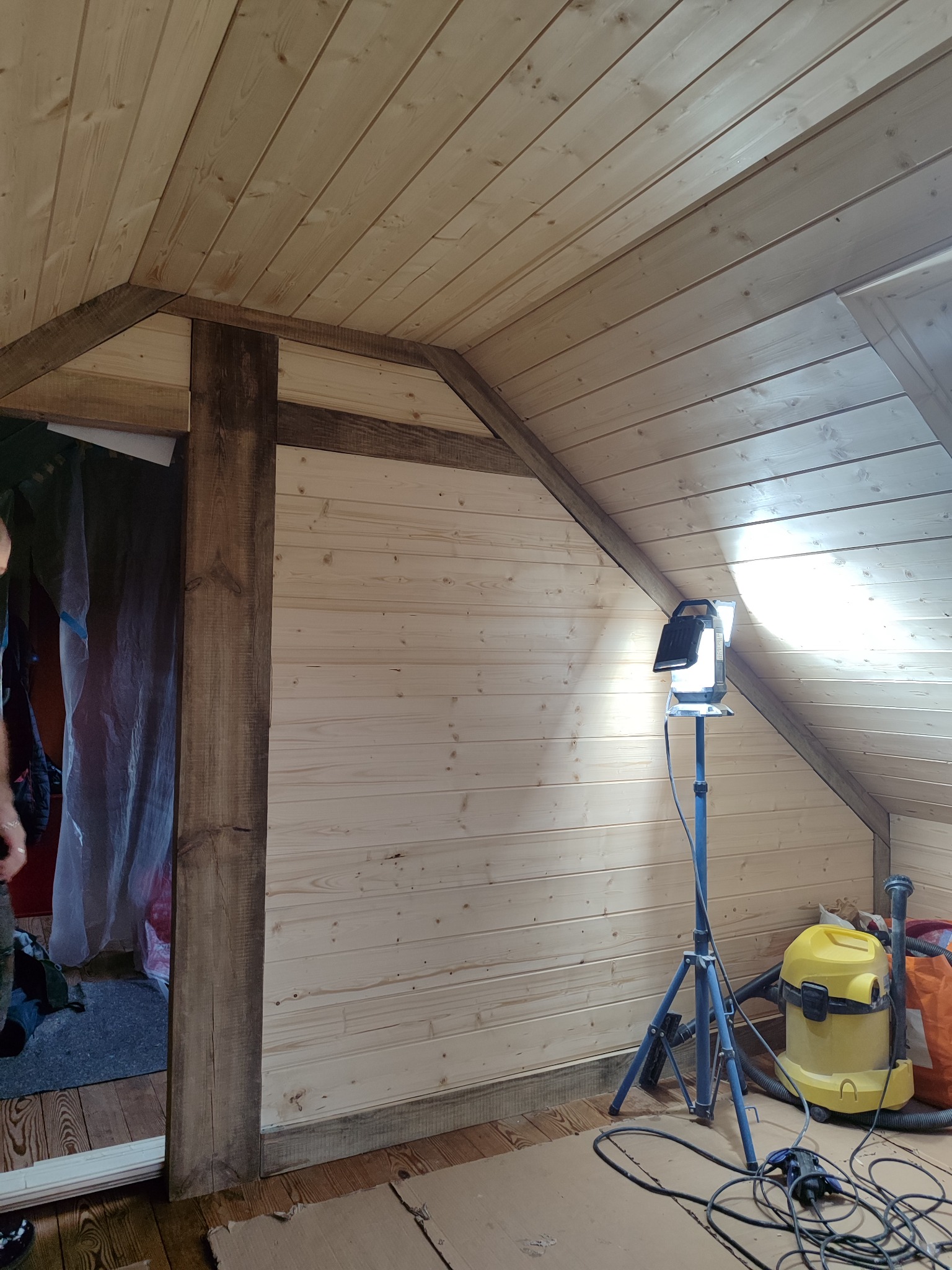 Poddasze wykończone jasną boazerią, z widoczną drewnianą konstrukcją nośną pomalowaną na ciemny brąz, oświetlone lampą budowlaną na statywie, z odkurzaczem przemysłowym w rogu i częściowo widoczną...