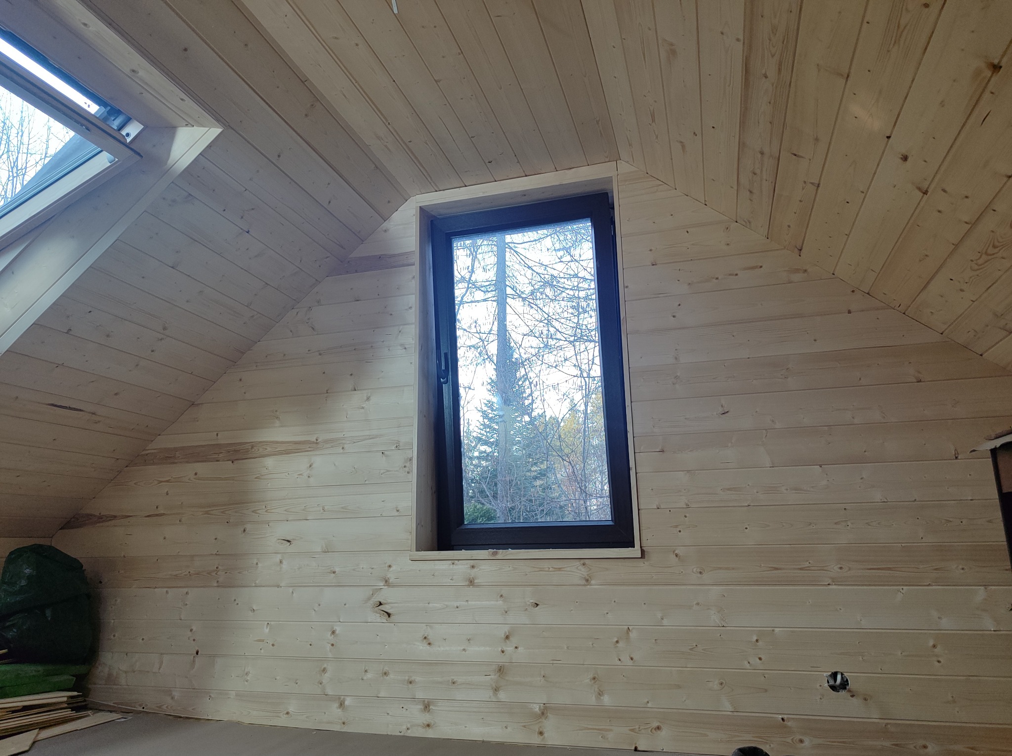 Poddasze wykończone jasną boazerią, z oknem dachowym i standardowym oknem z widokiem na drzewa, widoczny otwór na gniazdko elektryczne w ścianie.