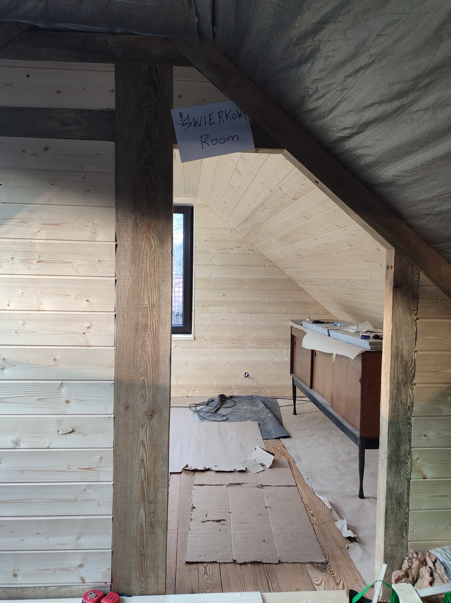 Poddasze w trakcie wykańczania boazerią, widoczne drewniane panele ścienne i sufitowe, stara komoda zabezpieczona folią, kartony na podłodze i prowizoryczny szyld z napisem 'Świerkowy Room'.