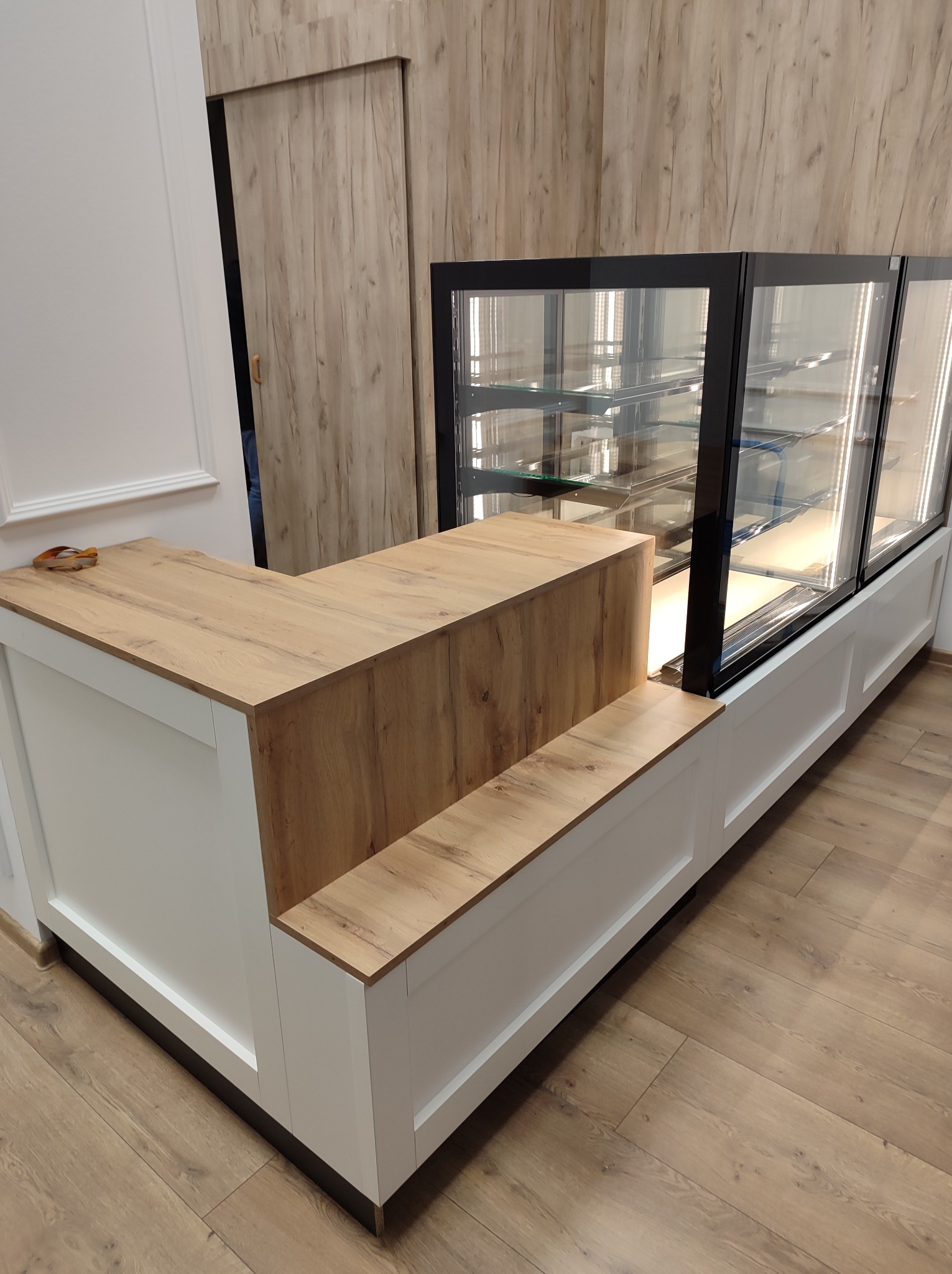 Nowoczesna lada recepcyjna z drewnianym blatem i witryną chłodniczą z oświetleniem LED, na podłodze panele imitujące drewno.