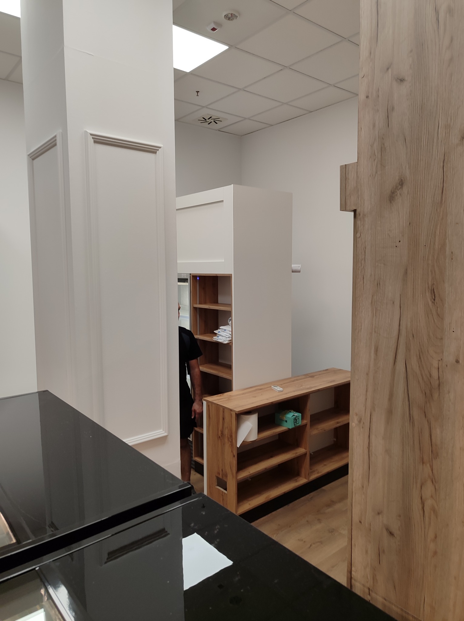 Montaż szafy wnękowej z drewnianymi półkami w jasnym pomieszczeniu, widoczny fragment czarnego blatu i częściowo zasłonięta osoba.