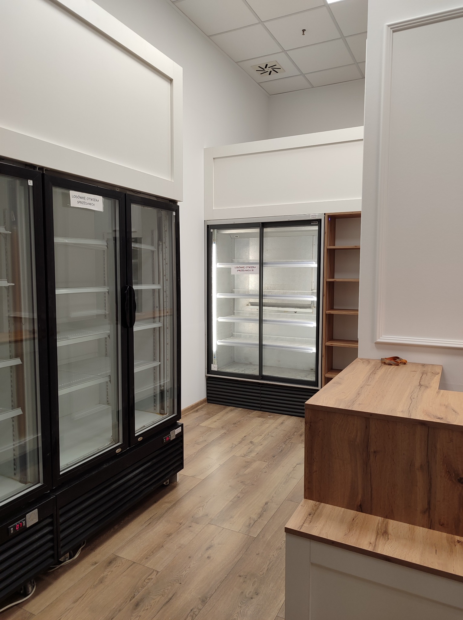 Wnętrze sklepu z pustymi chłodniami i drewnianą ladą, widoczne elementy wykończenia ścian.