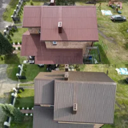 Widok z góry na dom z nowym, brązowym dachem z blachy trapezowej; widoczne elementy wykończeniowe komina i otoczenie domu z ogrodem, placem zabaw i zaparkowanym samochodem.
