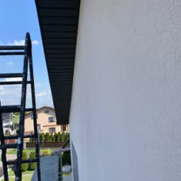 Świeżo pomalowana, jasna elewacja domu z widoczną drabina i fragmentem ciemnej podbitki dachowej na tle błękitnego nieba.